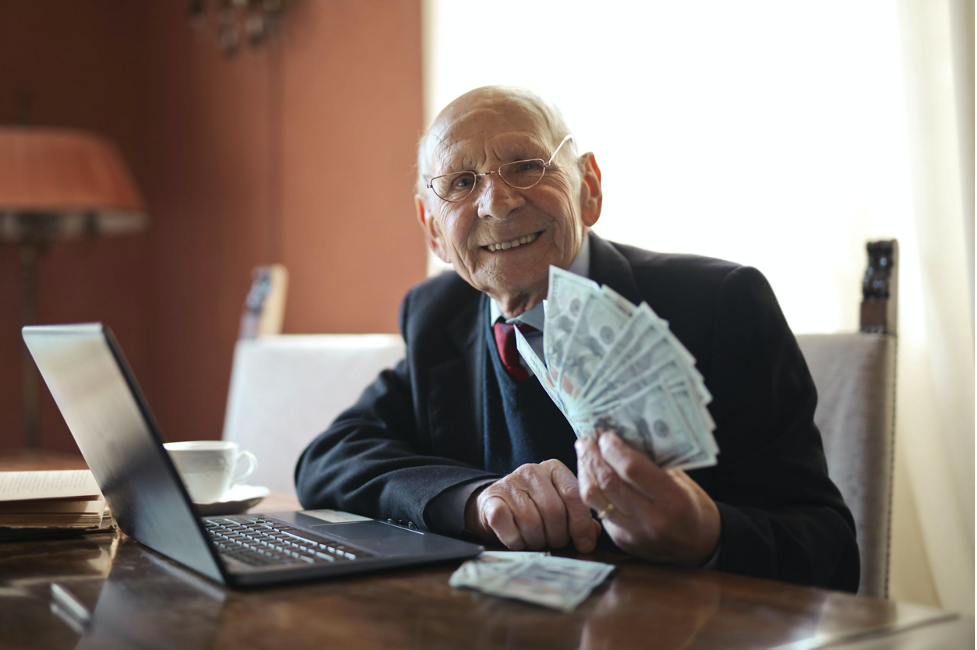 An old man, holding a stash of cash infront of a laptop